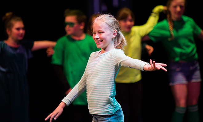  A young girl performing on stage in spotlight while other performers watch. 