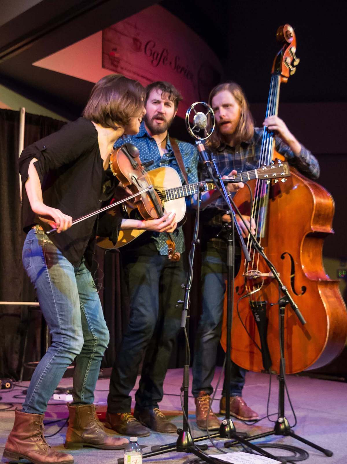  Three musicians playing a guitar, fiddle and bass harmonizing around one mic. 